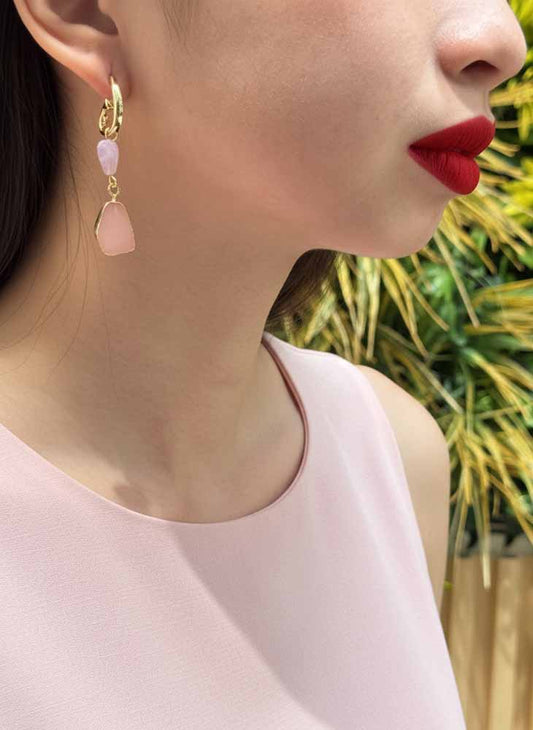 Close-up of a person wearing gold earrings with pink stones, red lipstick, and a light pink top against a blurred natural background.