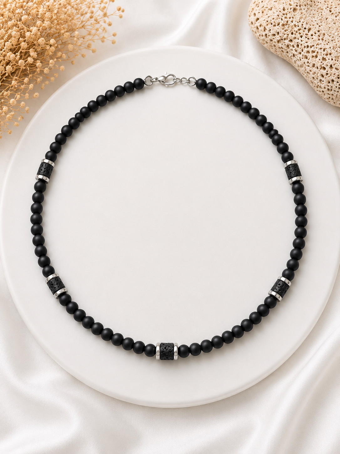 A black beaded necklace with silver-toned spacer accents and a silver clasp, displayed on a round white plate with decorative dried flowers and textured elements in the corners.