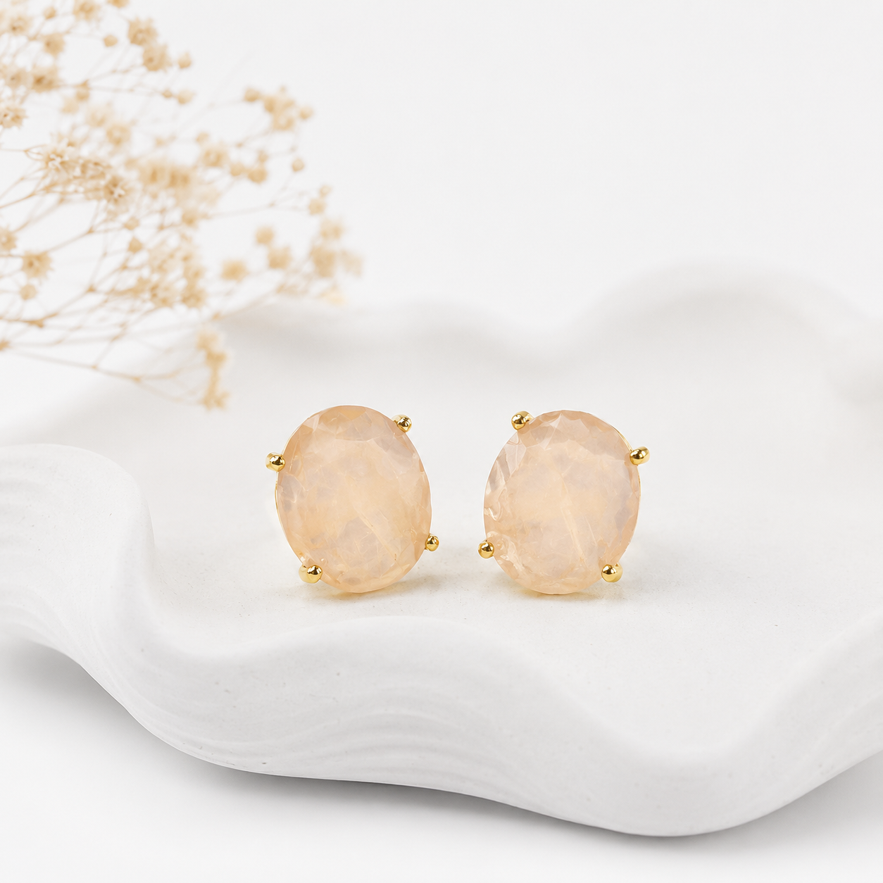 A pair of oval stud earrings with pale peach-pink faceted gemstones in gold prong settings, displayed on a white wavy ceramic dish with dried baby's breath flowers in the background.