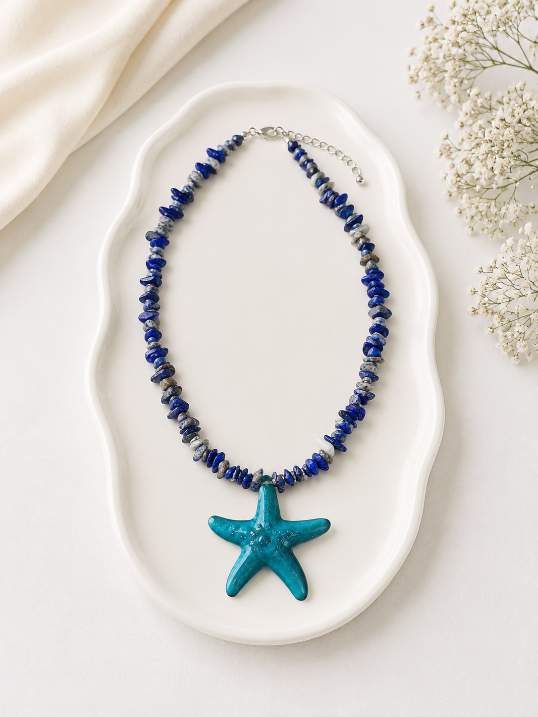 Necklace with blue beads and starfish pendant on a white dish with flowers in the background