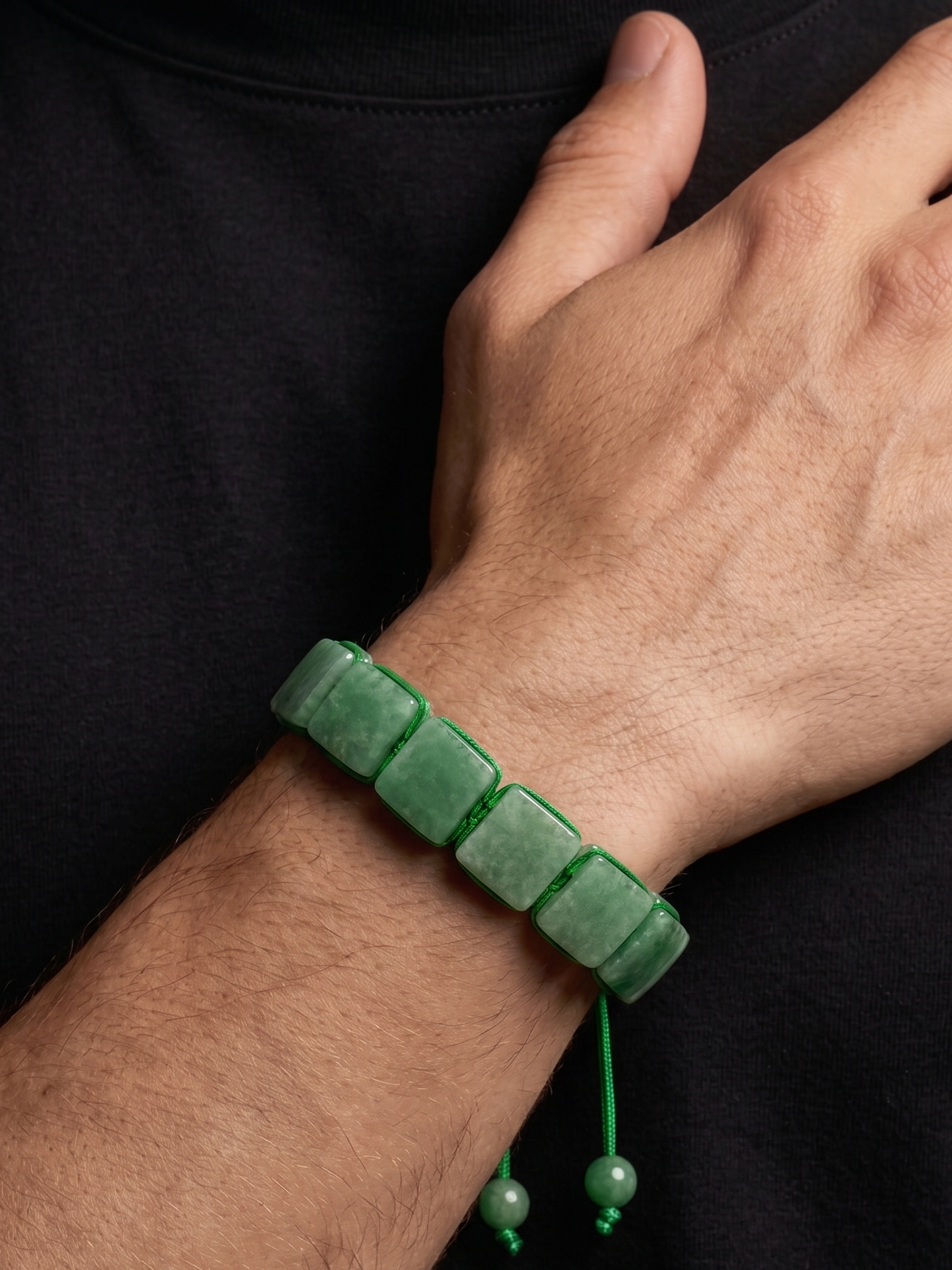 A green beaded bracelet with a cord and dangling beads, worn on a person's wrist against a black shirt background.