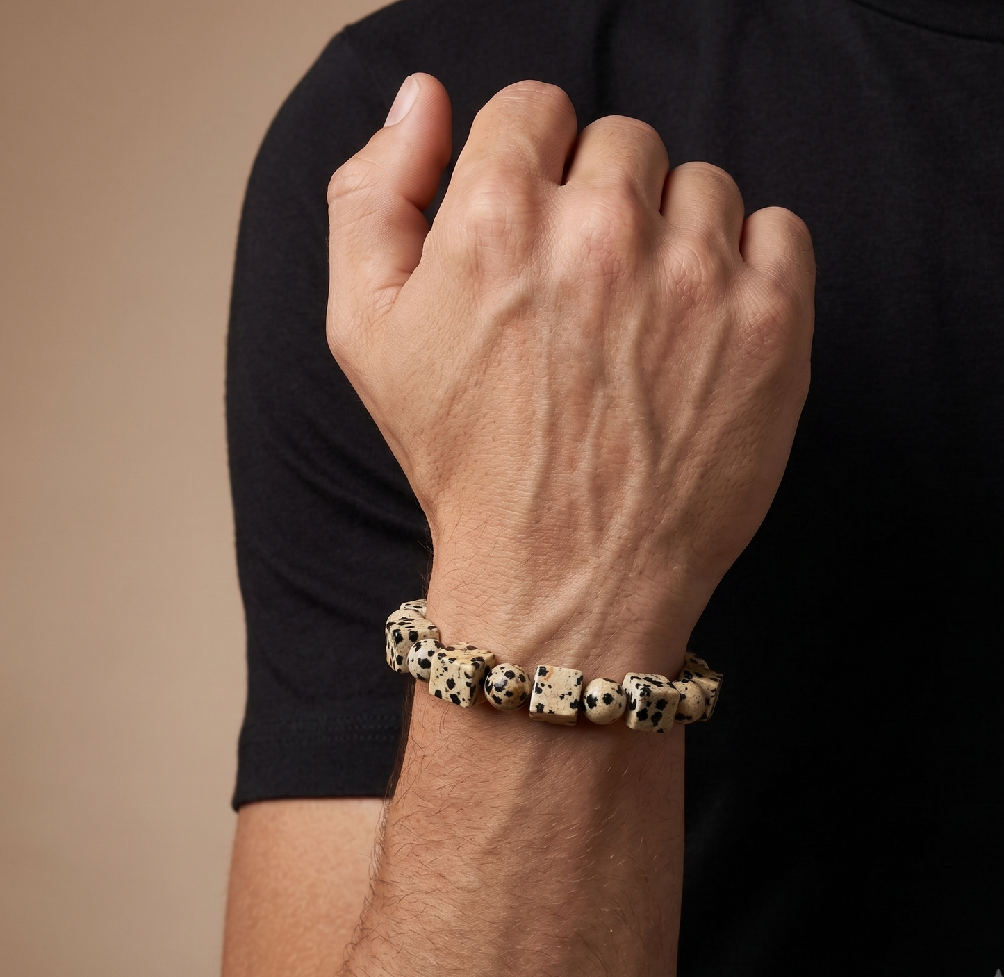 A close-up of a person's hand and forearm wearing a beaded bracelet made of cream-colored beads with black spots, resembling dalmatian spots, set against a black t-shirt and a warm beige background.