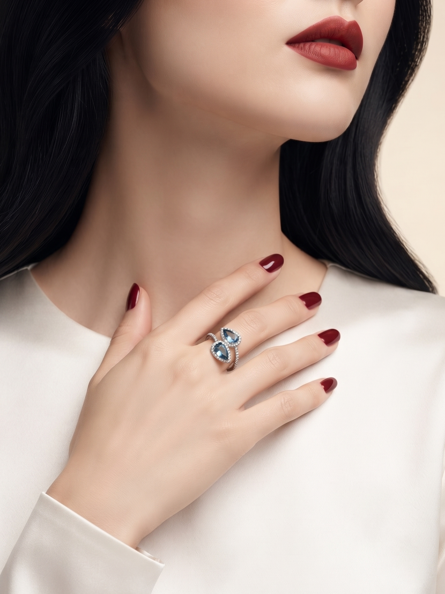A close-up of a woman wearing a silver ring with a four-petal clover design set with light blue gemstones, styled with dark red nail polish and a white satin top.
