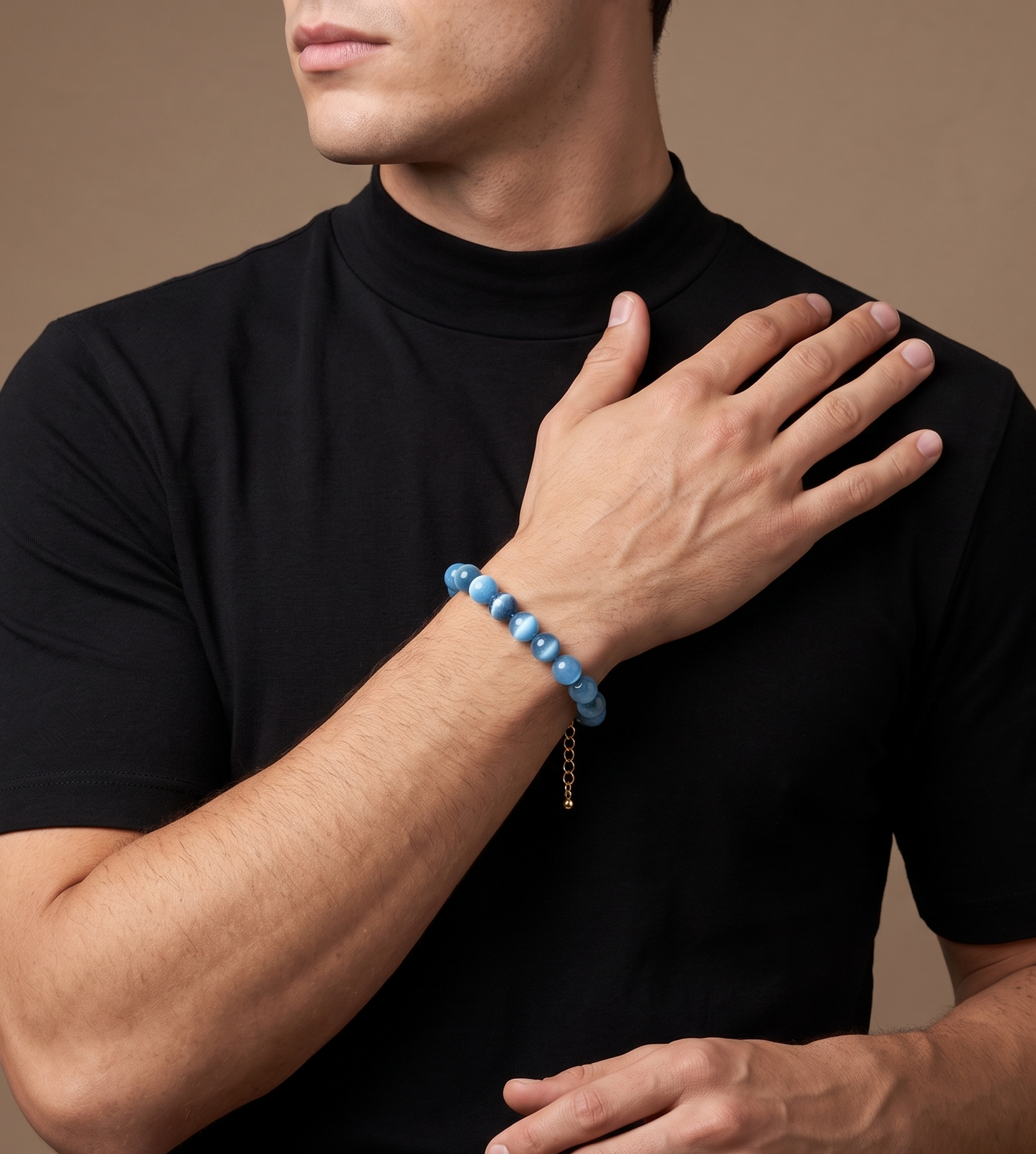 A man wearing a black mock-neck t-shirt and a light blue beaded bracelet with a small gold chain accent, posed against a solid warm brown background.
