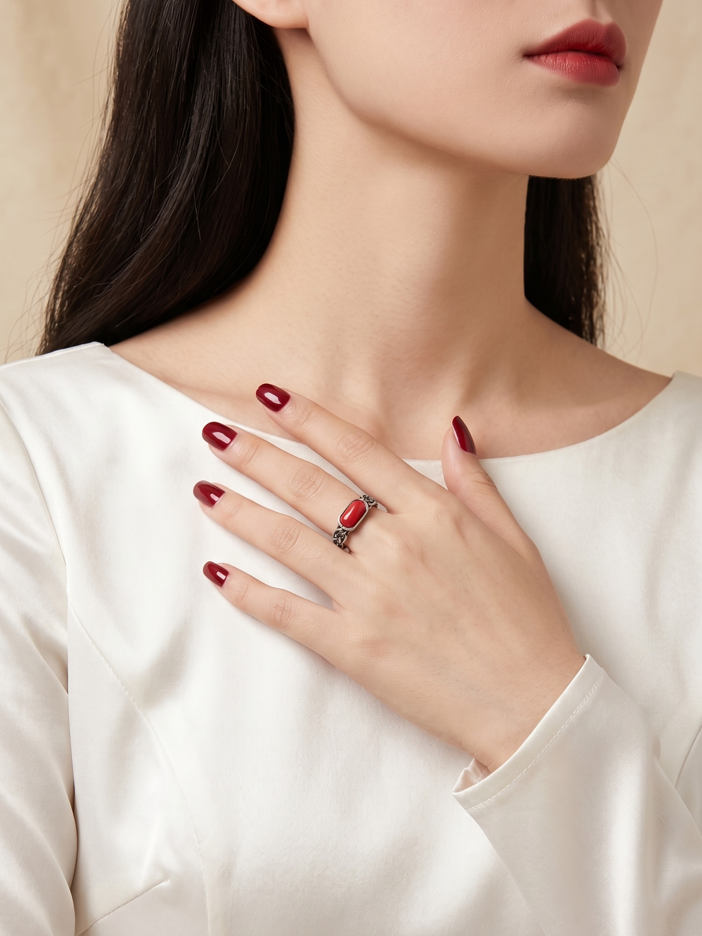 A close-up of a woman wearing a silver ring with a red oval stone, styled with a white satin top and dark red nail polish against a soft beige background.