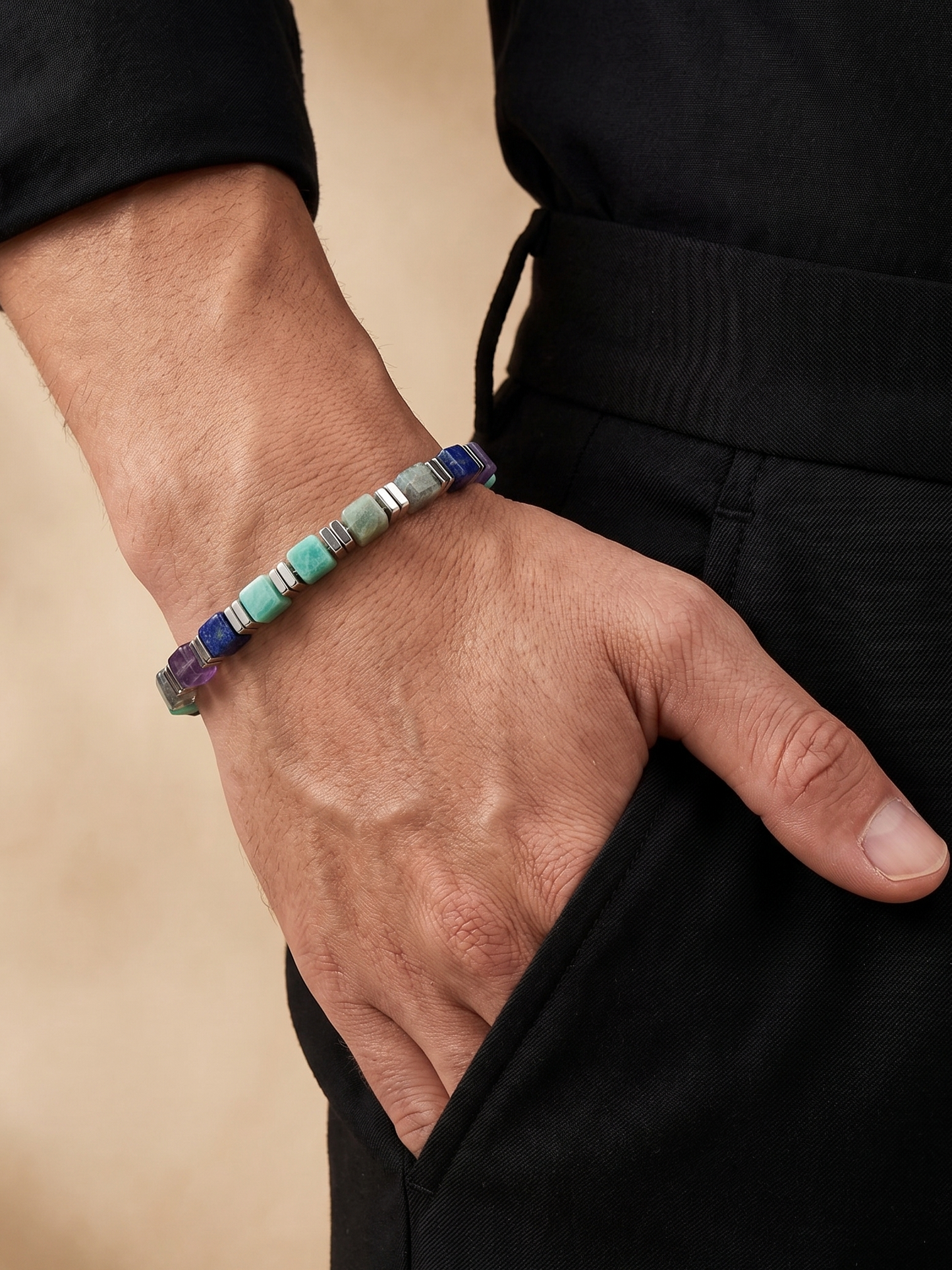 A close-up of a person's hand wearing a beaded bracelet made of colorful stone beads, paired with a black long-sleeve top and black trousers against a warm beige background.