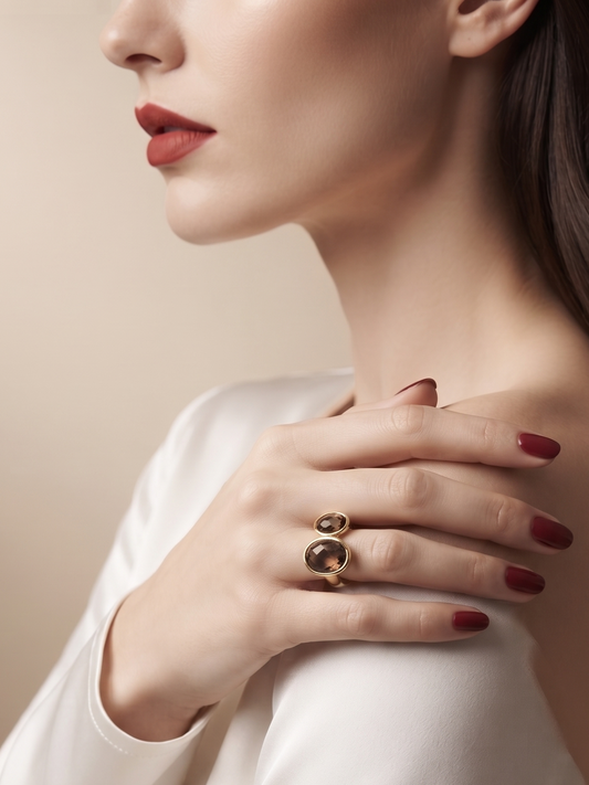 A close-up lifestyle shot of a model wearing a gold ring with two interlocking oval shapes, styled with a white satin top against a warm beige background.