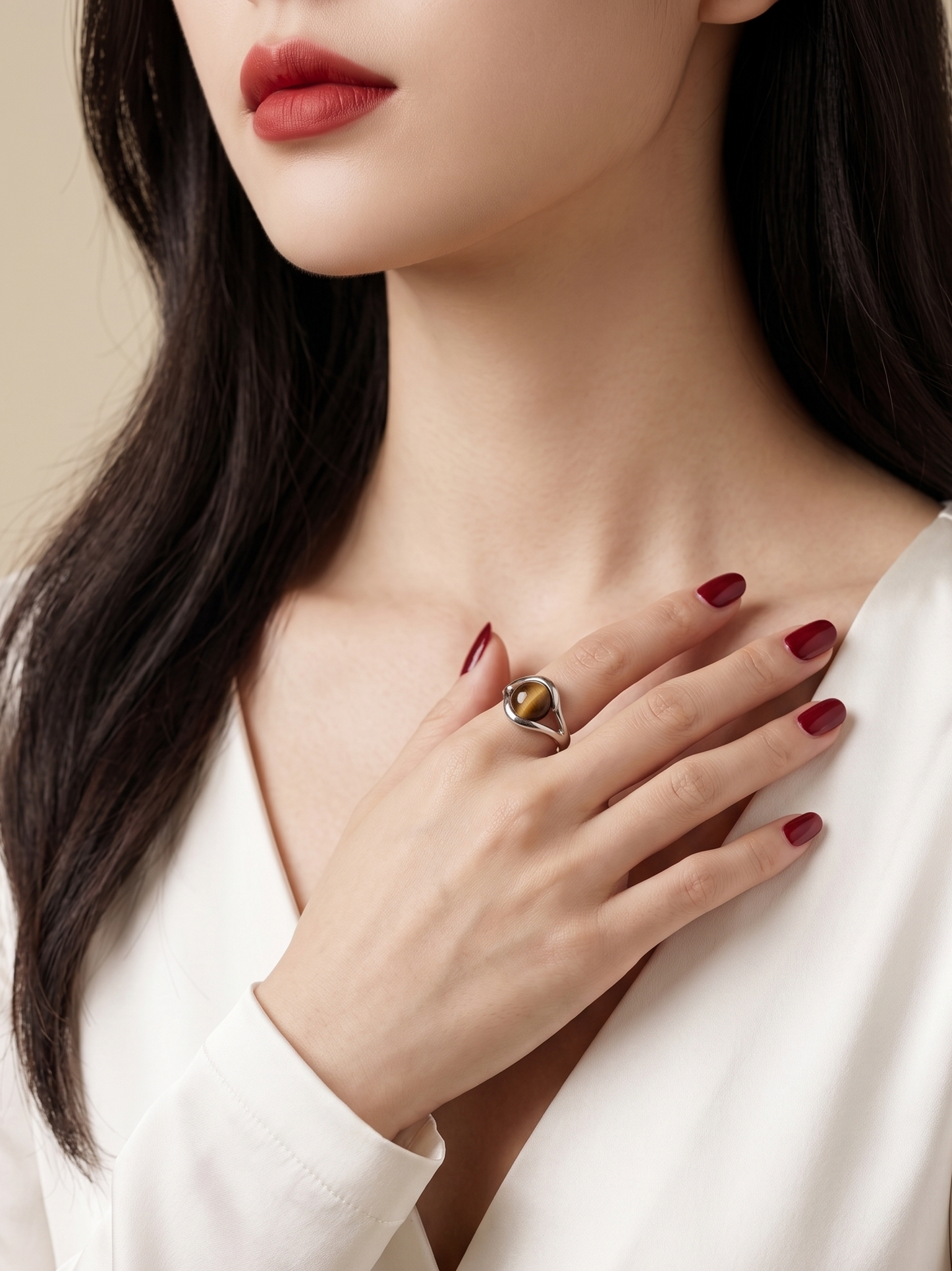 A close-up of a woman wearing a silver-toned ring with a gold and black triangular stone, styled with a white satin top and dark red nail polish.