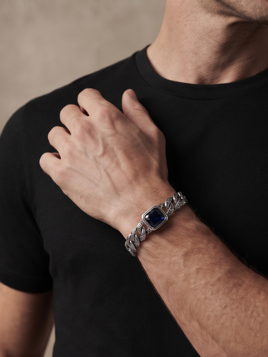 A close-up of a man wearing a silver-toned chain-link bracelet with a square dark blue bezel on his wrist, paired with a black t-shirt.
