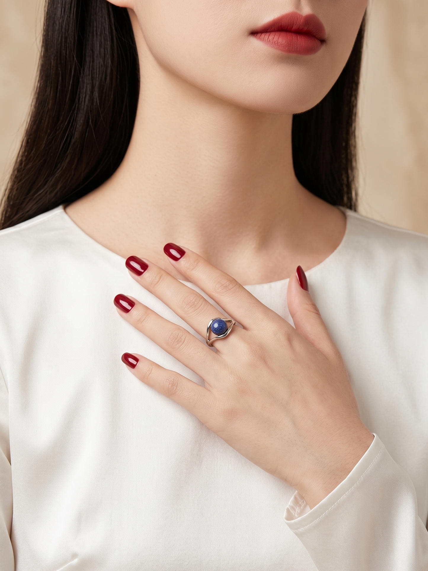 A close-up of a woman wearing a silver ring with a round blue gemstone, styled with a white satin top and dark red manicured nails.