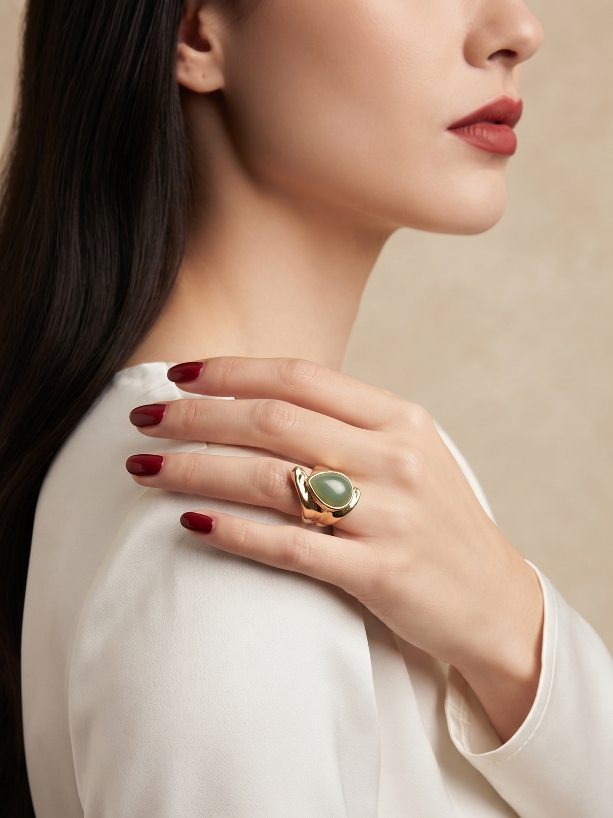 A close-up lifestyle shot of a model wearing a gold ring with a green teardrop-shaped stone, set against a soft beige background.