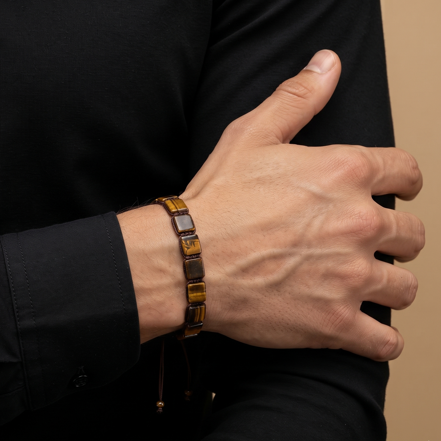 A close-up of a person's hand and forearm wearing a brown leather bracelet with polished gold-toned rectangular links and dark square accents, set against a black shirt and a warm beige background.