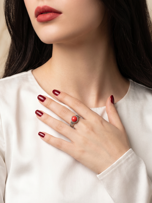 A close-up lifestyle shot of a model wearing a silver ring with a round red gemstone, styled with dark red nail polish and a white satin top.