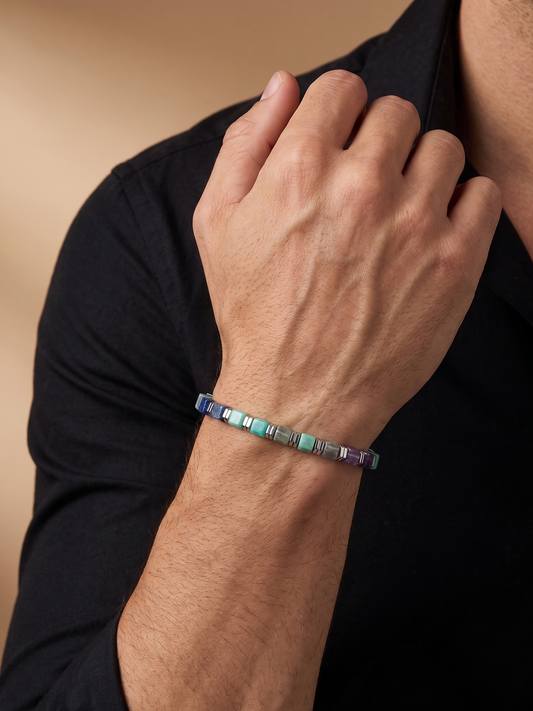 A close-up of a person's hand and wrist wearing a beaded bracelet with iridescent square beads in shades of blue, green, and purple, worn over a black shirt.