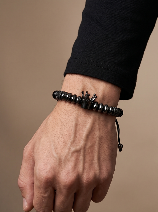 A close-up of a man's hand and wrist wearing a black beaded bracelet with a small crown charm, set against a solid warm beige background.