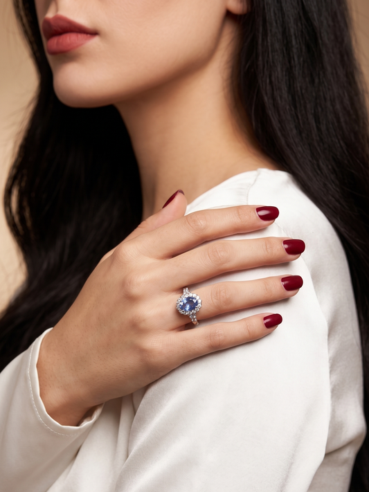 A close-up lifestyle shot of a woman wearing a silver ring with a round blue gemstone on her finger, resting her hand on her shoulder. She is wearing a white top against a warm beige background.