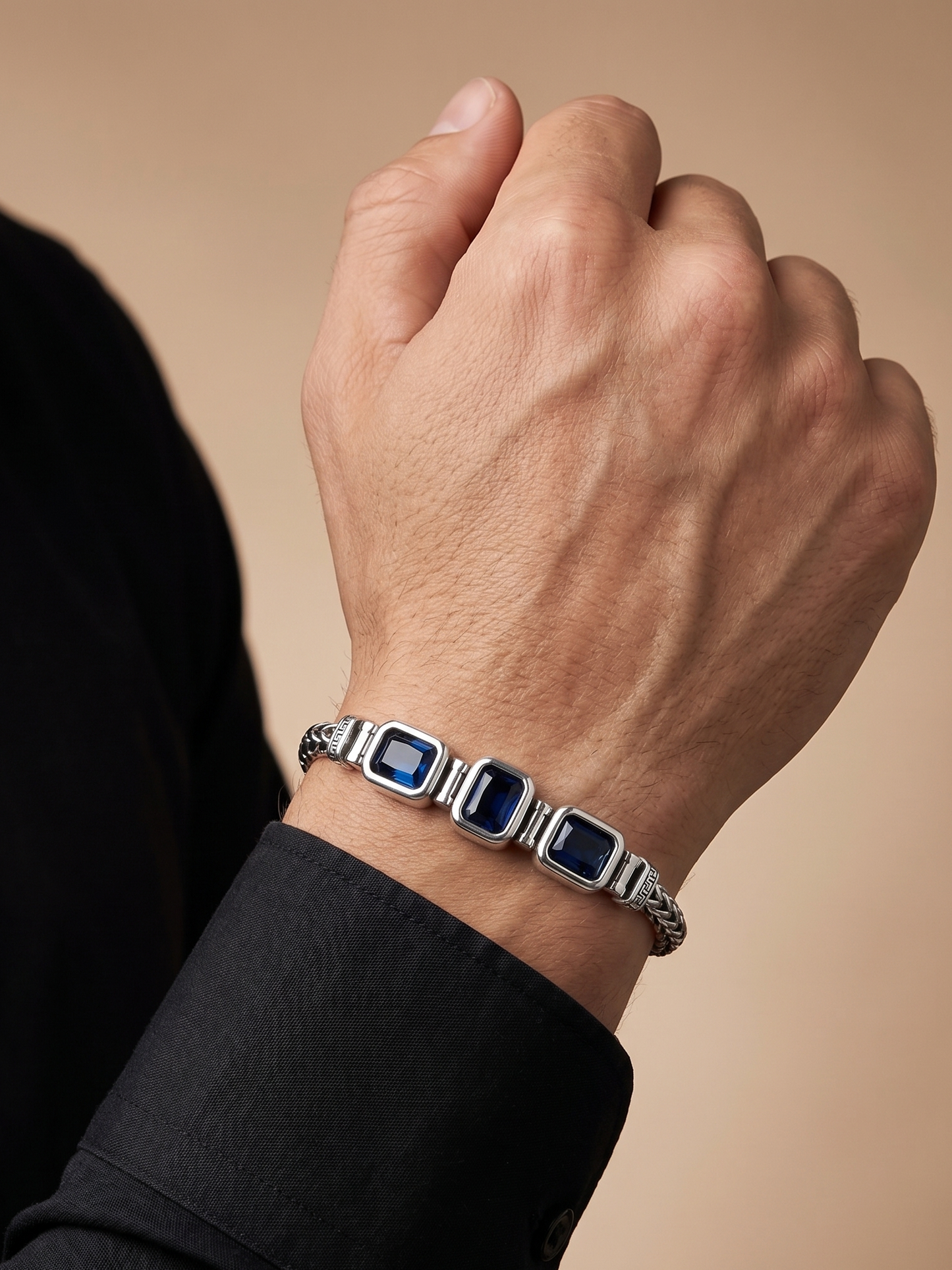 A close-up of a man's wrist wearing a silver-toned bracelet with three rectangular blue gemstones, set against a warm beige background.