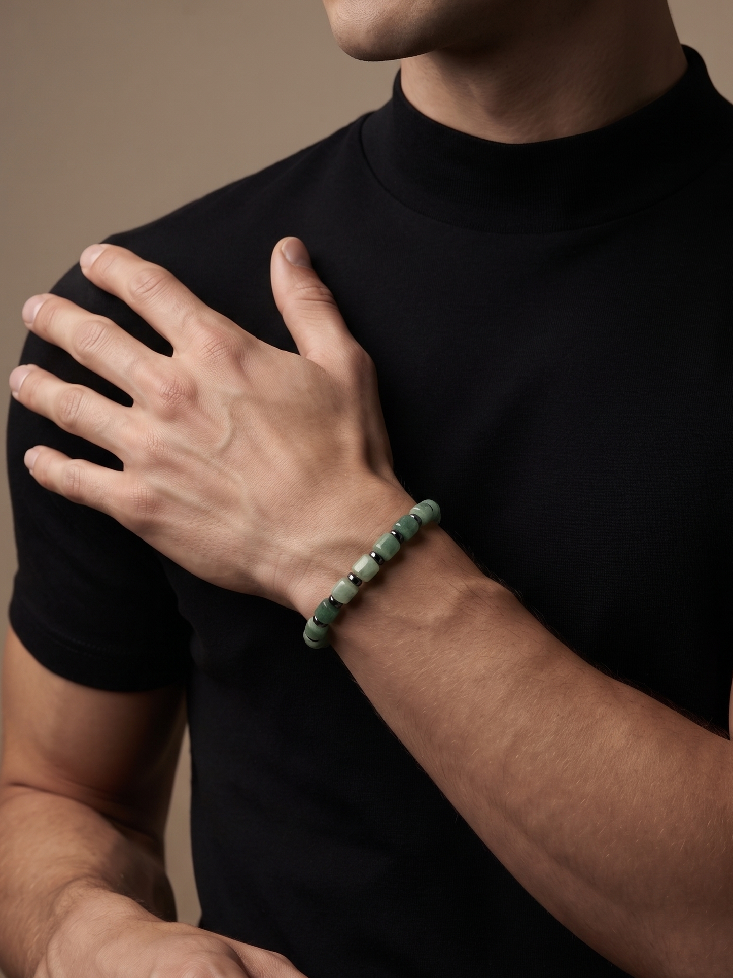 A close-up of a man wearing a black t-shirt and a green beaded bracelet on his wrist, with his hand resting on his chest against a solid beige background.