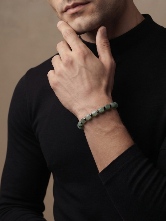 A man wearing a black long-sleeve mock neck shirt and a green beaded bracelet, posing with his hand on his chin against a textured beige background.