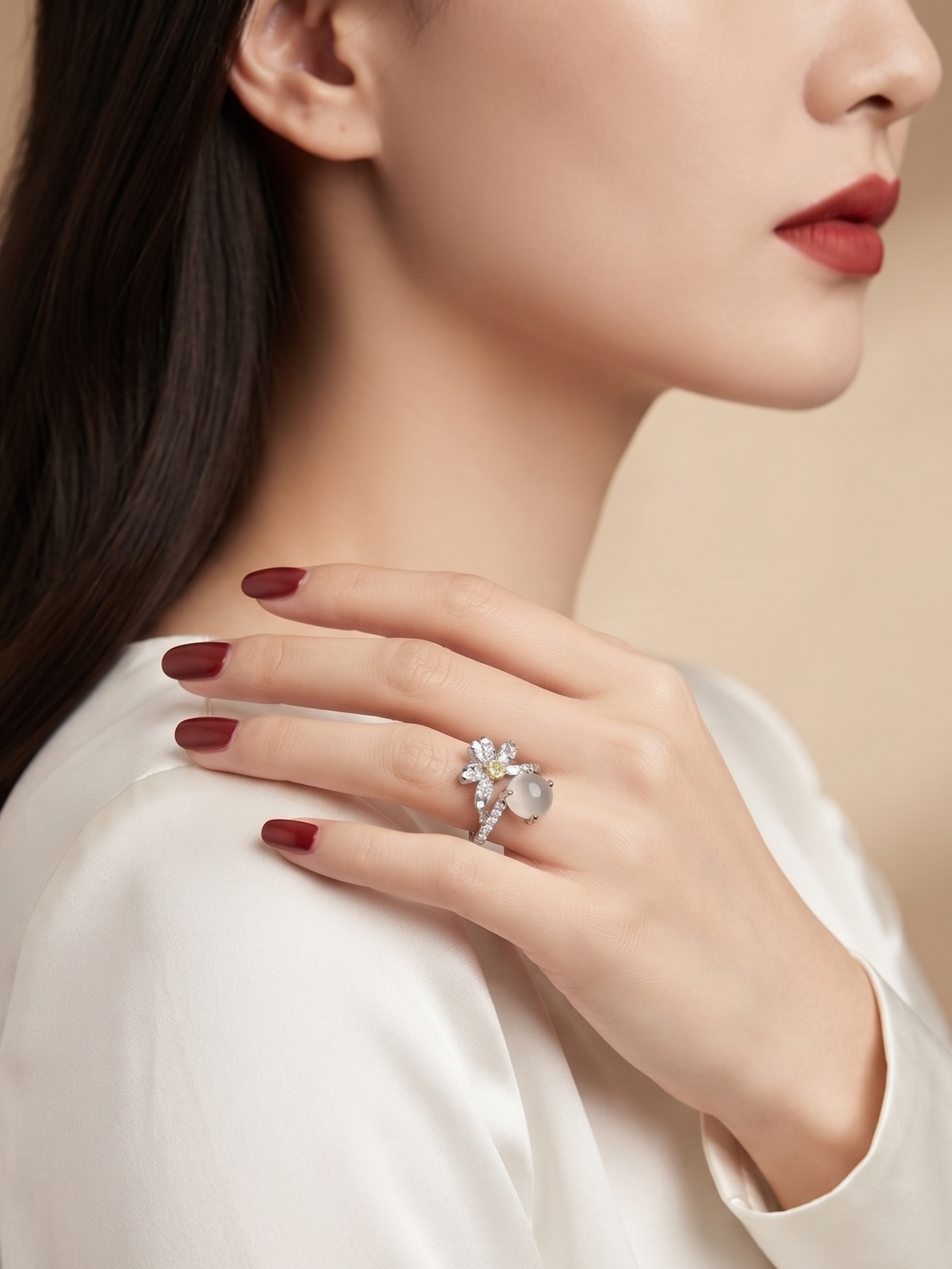A close-up lifestyle shot of a woman wearing a silver-toned flower ring with clear stones, styled with dark red nail polish and a white satin top against a warm beige background.
