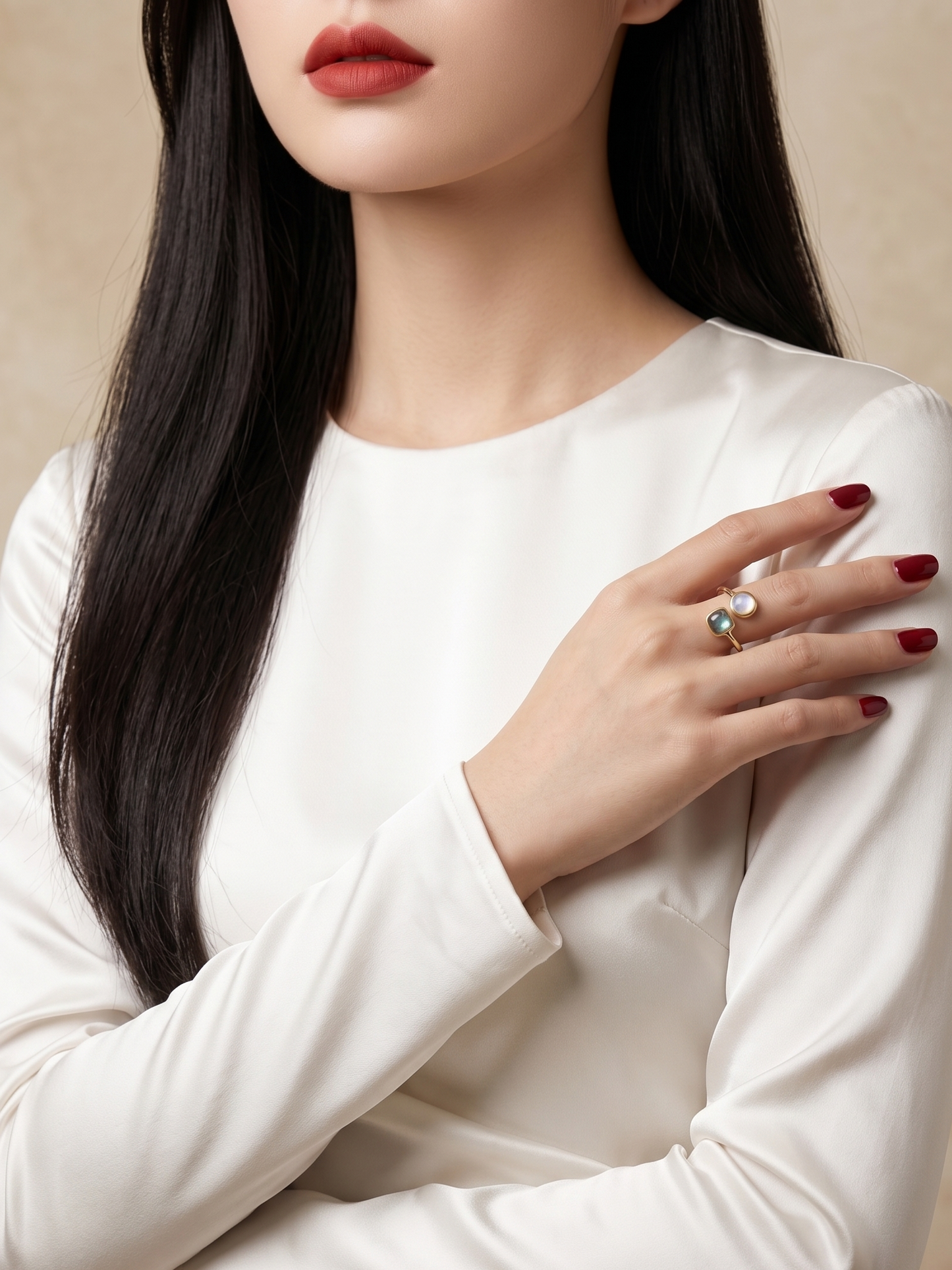 A close-up of a woman wearing a white long-sleeve top and a silver ring with a white pearl and green stone, posed against a warm beige background.