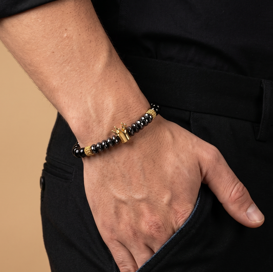 A close-up of a man's hand in his pocket wearing a beaded bracelet with black and gold beads and a gold cross charm, dressed in a black shirt and trousers against a beige background.