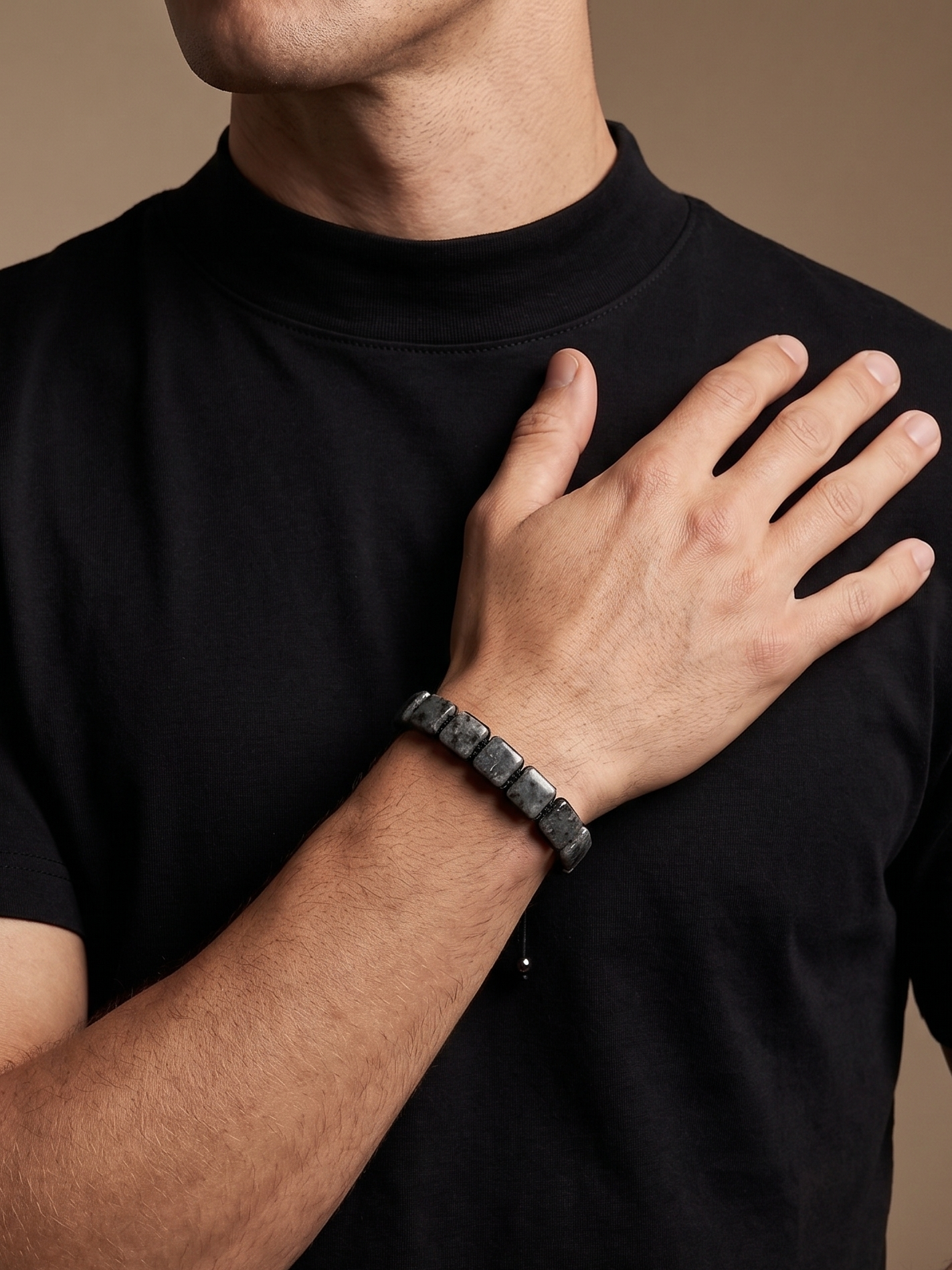 A close-up of a man wearing a black short-sleeve mock neck t-shirt and a dark gray beaded bracelet on his wrist, set against a solid warm beige background.