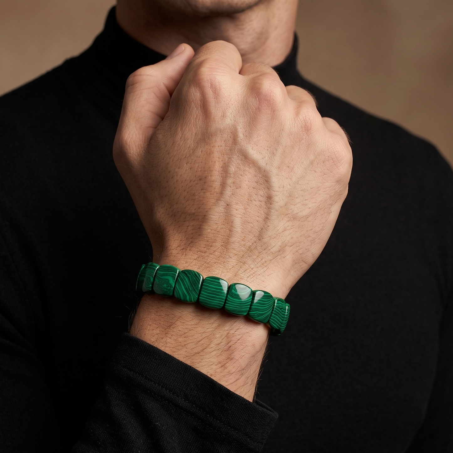 A close-up of a man's wrist wearing a green faceted stone bracelet, shown against a black turtleneck sweater and a warm brown background.