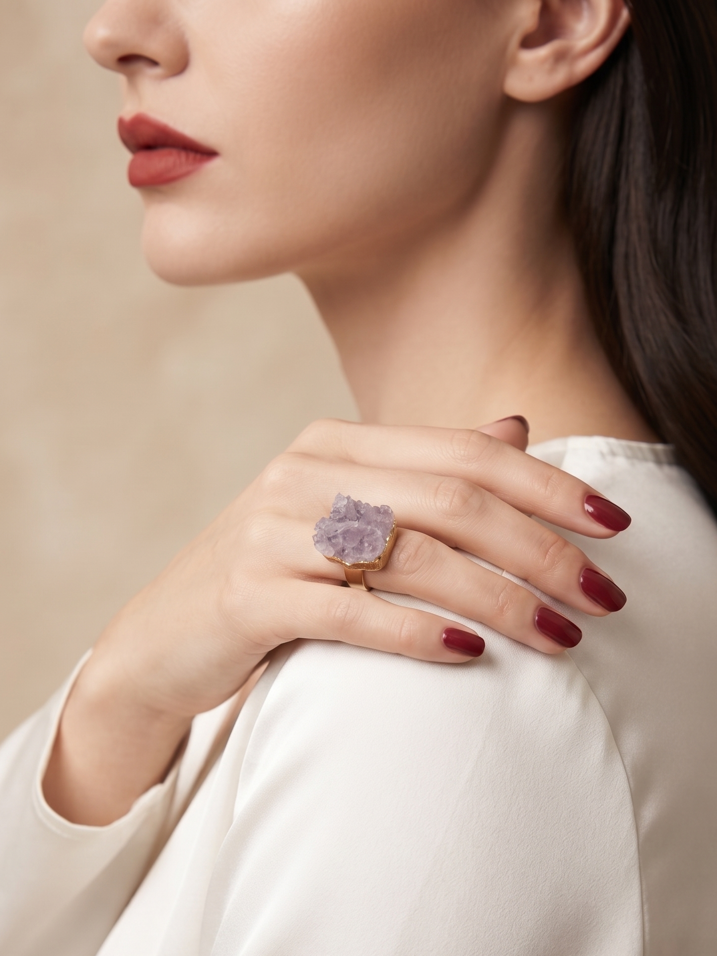 A close-up lifestyle shot of a woman wearing a chunky ring with a lavender crystal cluster on a gold band, set against a soft beige background.