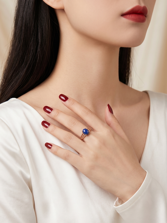 A close-up lifestyle shot of a woman wearing a delicate rose gold ring with a round blue gemstone, resting her hand on her chest. She is wearing a white satin top, and the background is a soft, warm beige tone.