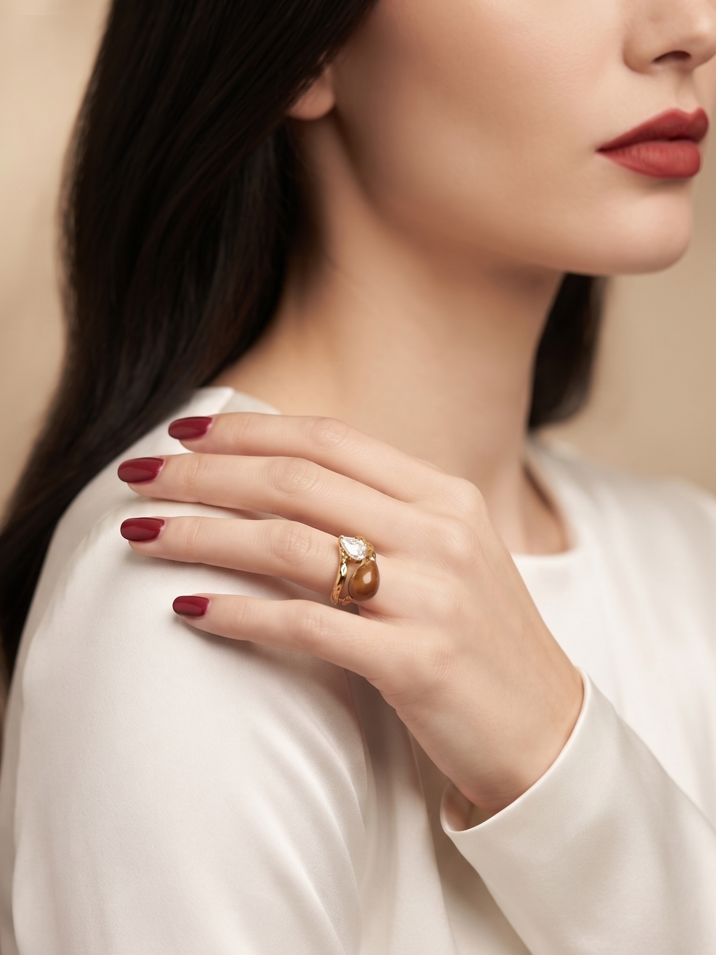 A close-up lifestyle shot of a woman wearing a gold ring with a clear gemstone on her finger, resting her hand on her shoulder. She is wearing a white satin top against a soft beige background.