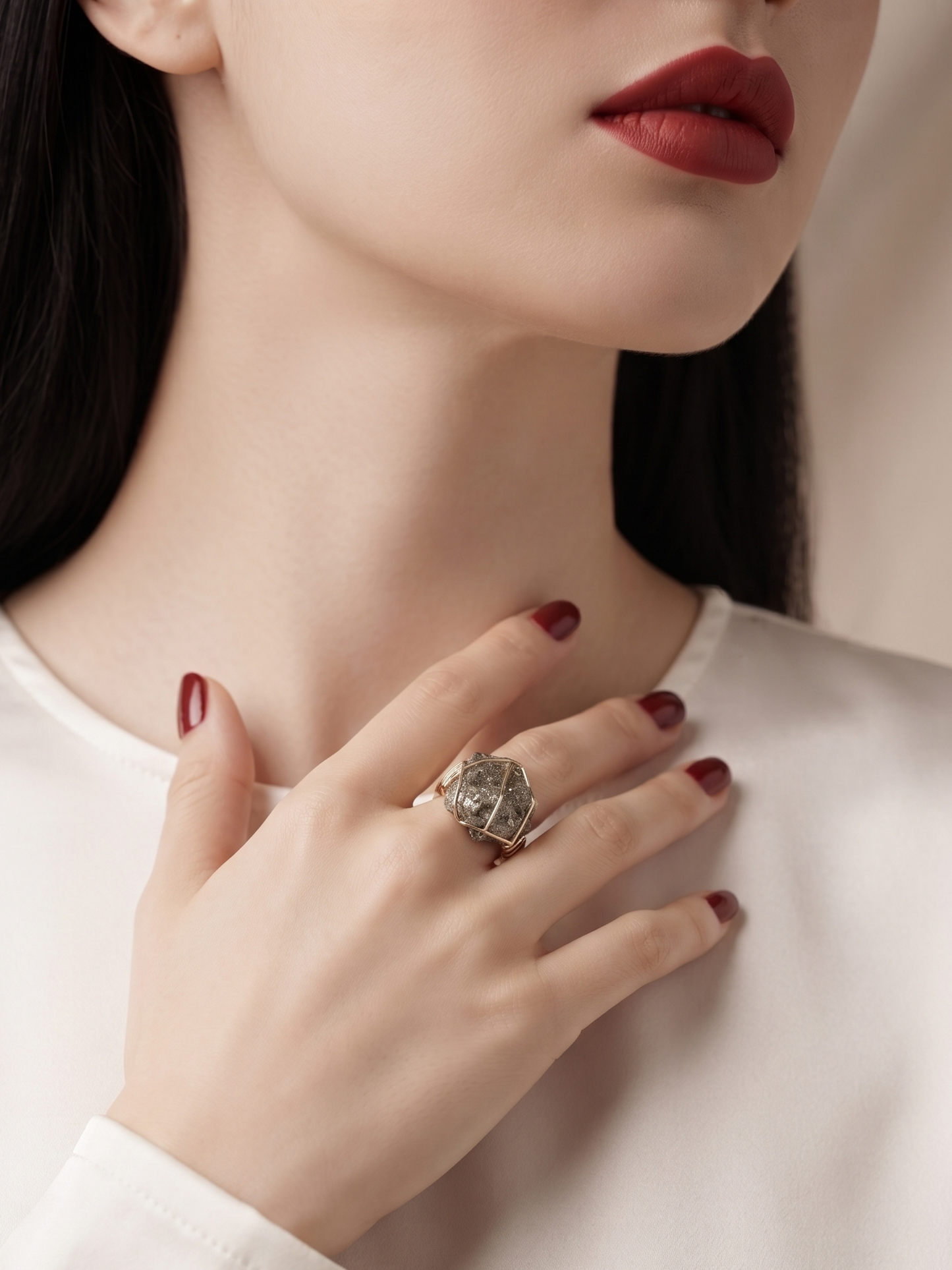 A close-up lifestyle shot of a woman wearing a large, ornate silver ring with a detailed floral or scrollwork design on her ring finger. She is wearing a white satin long-sleeve top, dark red lipstick, and dark red nail polish, with her hand resting on her chest against a soft beige background.