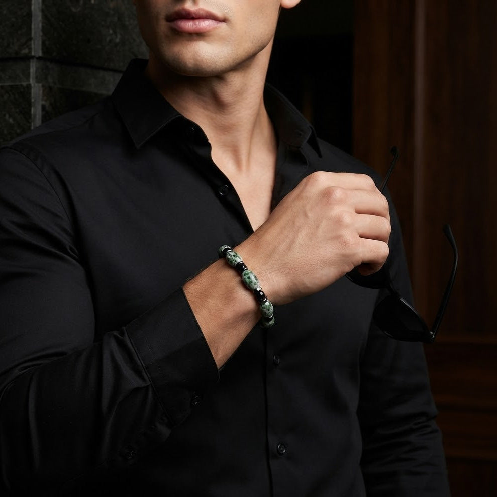 A man wearing a black button-down shirt and a green beaded bracelet, holding a pair of sunglasses in a dimly lit indoor setting.