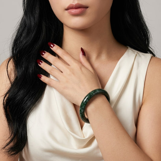 A close-up of a woman wearing a dark green jadeite bangle bracelet, with her hand resting on her chest. She is wearing a cream-colored satin top, and her dark hair and red manicured nails are visible against a neutral gray background.