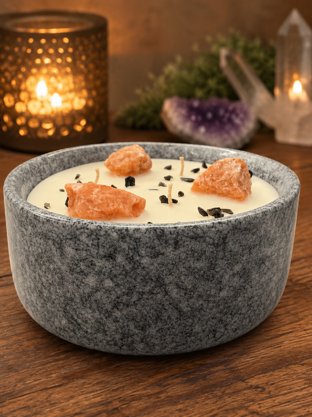 Candle in a gray stone bowl with decorative elements on a wooden surface, surrounded by candles and crystals.