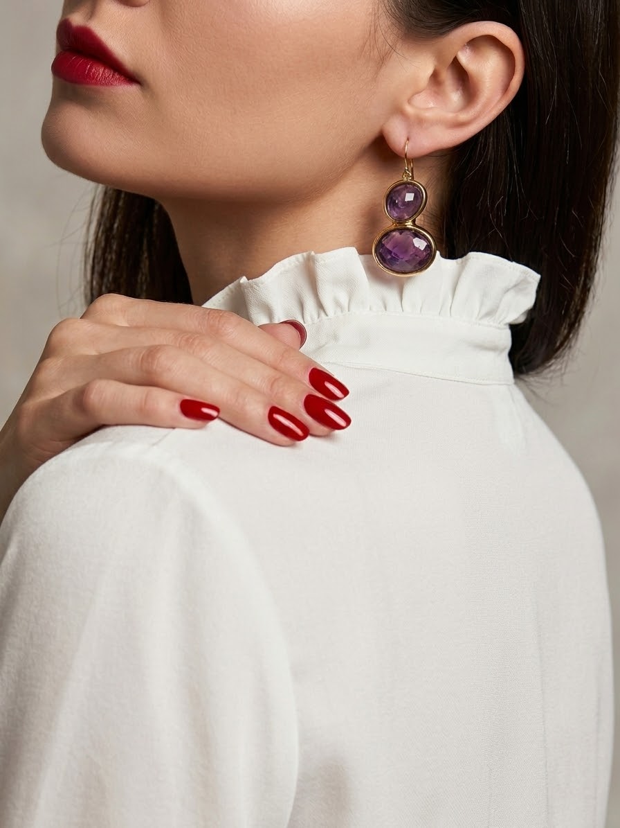 A close-up of a woman wearing a pair of double-stone dangle earrings with purple amethyst gemstones in gold settings, paired with a white ruffled mock neck top.