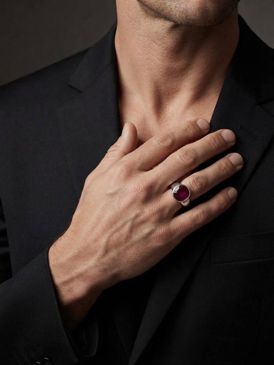 A close-up of a man in a black suit wearing a silver ring with a round deep red gemstone on his hand.