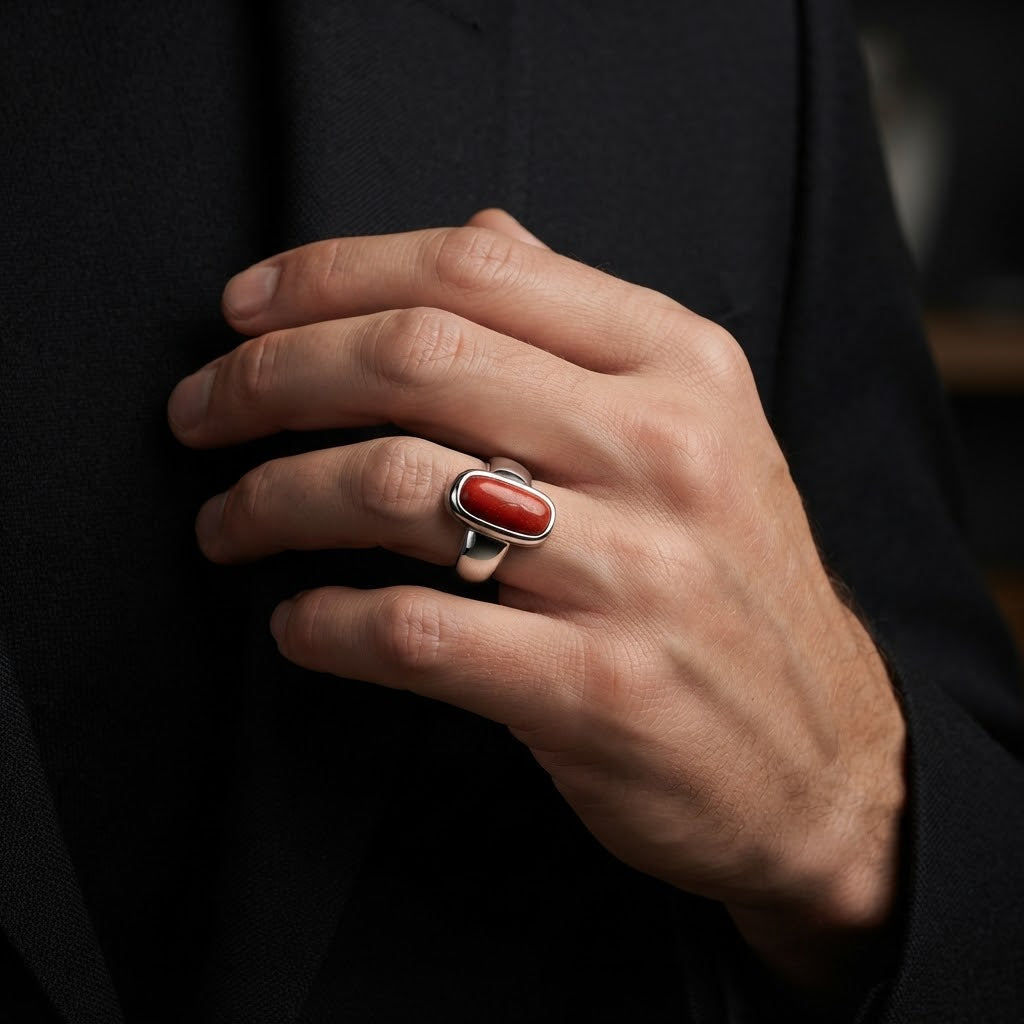 A close-up of a hand wearing a silver ring with an oval red cabochon stone, set against a dark black suit jacket.