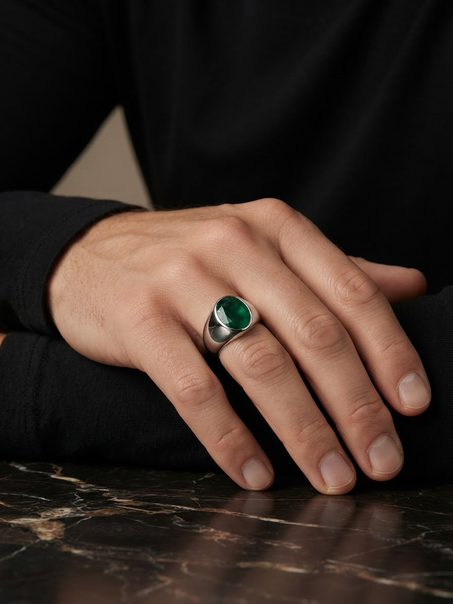 A close-up of a hand wearing a silver ring with a round green gemstone, resting on a dark marble surface against a black background.
