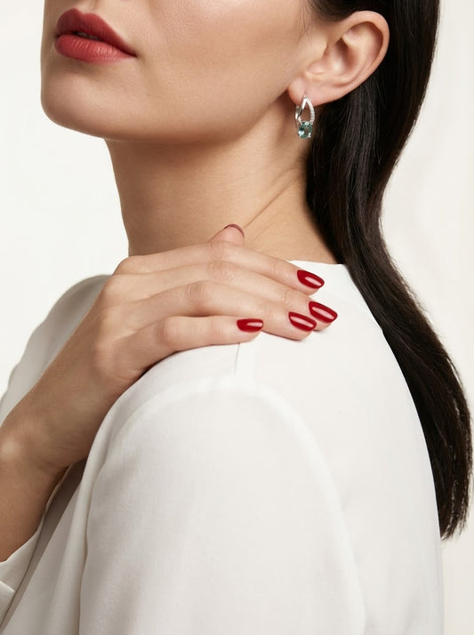Close-up of a woman wearing a small silver crescent-shaped hoop earring with a light blue stone, paired with a white top and red nail polish.