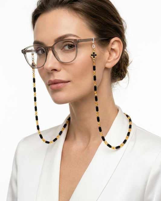 A woman wearing rectangular eyeglasses with long dangling chain straps made of alternating black and gold beads, shown against a clean white background.
