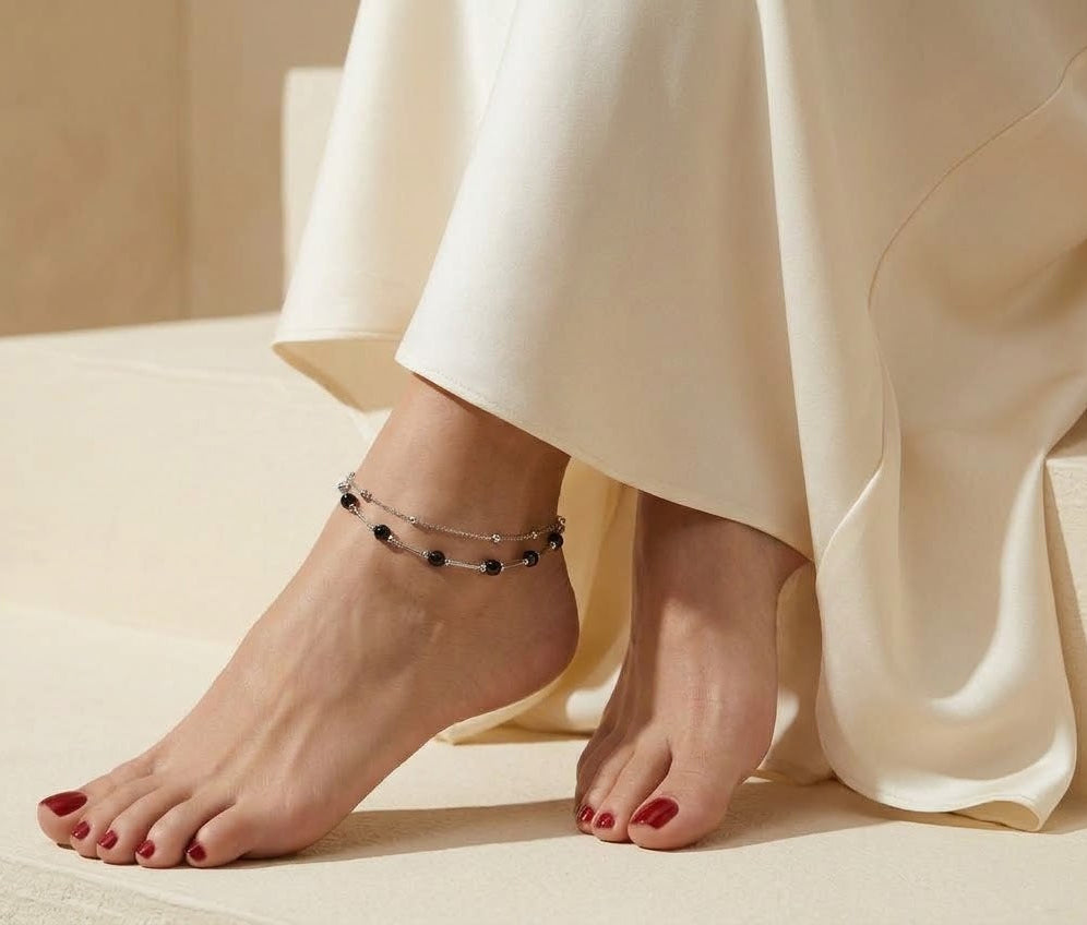 A close-up lifestyle shot of a person's bare feet and ankles wearing a delicate silver anklet with small dark beads, styled with a cream satin dress on a beige stone staircase.