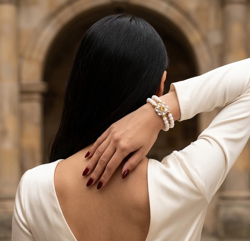 A woman with long black hair is shown from behind, wearing a cream-colored open-back top and a multi-strand pearl bracelet with gold floral accents on her wrist, posed in front of a stone archway.