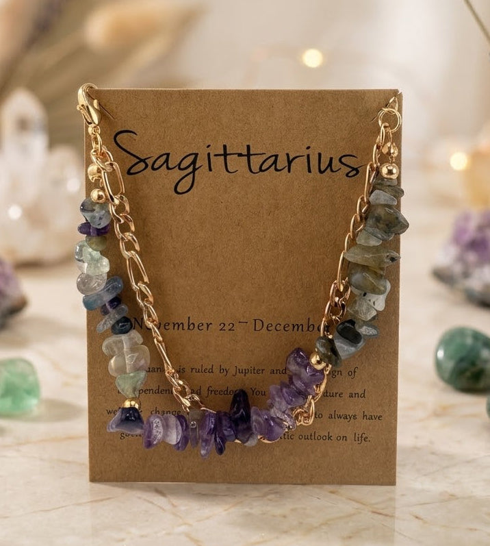 A gold chain necklace with multi-strand beads and purple amethyst chip accents, displayed on a brown kraft card labeled 'Sagittarius' in a styled setting with crystals and dried lavender.