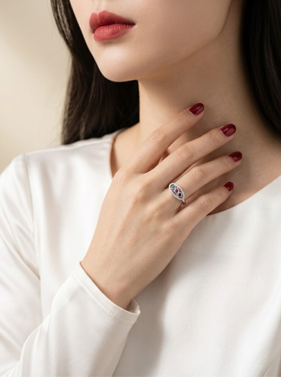 A close-up lifestyle shot of a woman wearing a silver ring with a heart-shaped blue and red gemstone cluster, styled with a white long-sleeve top and dark red nail polish.