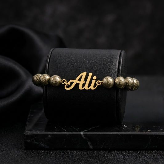 Gold bracelet with 'Ali' charm on a black box with 'Ali' branding, dark background