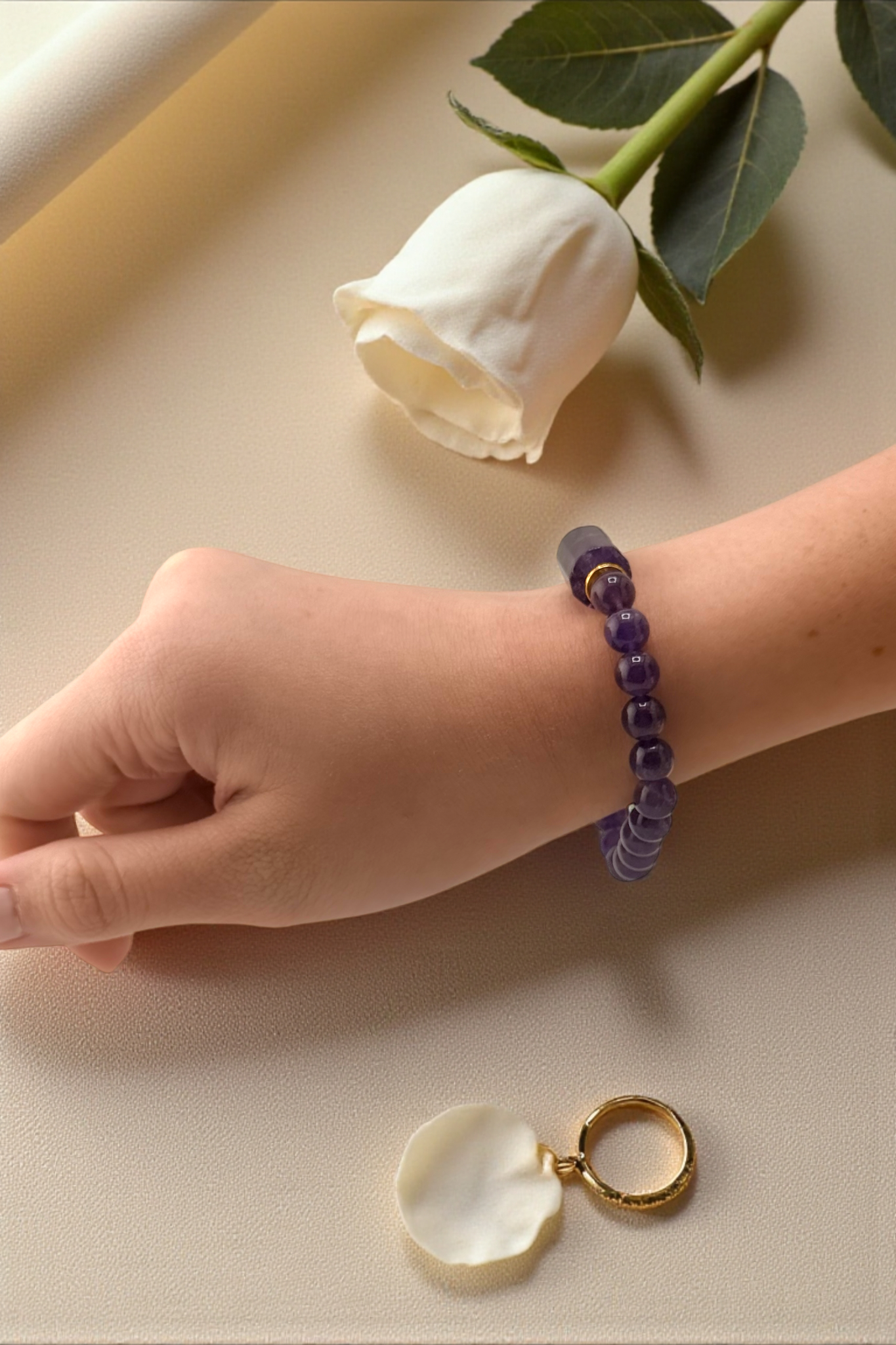 Hand wearing a purple beaded bracelet with a white rose and gold ring on a beige surface