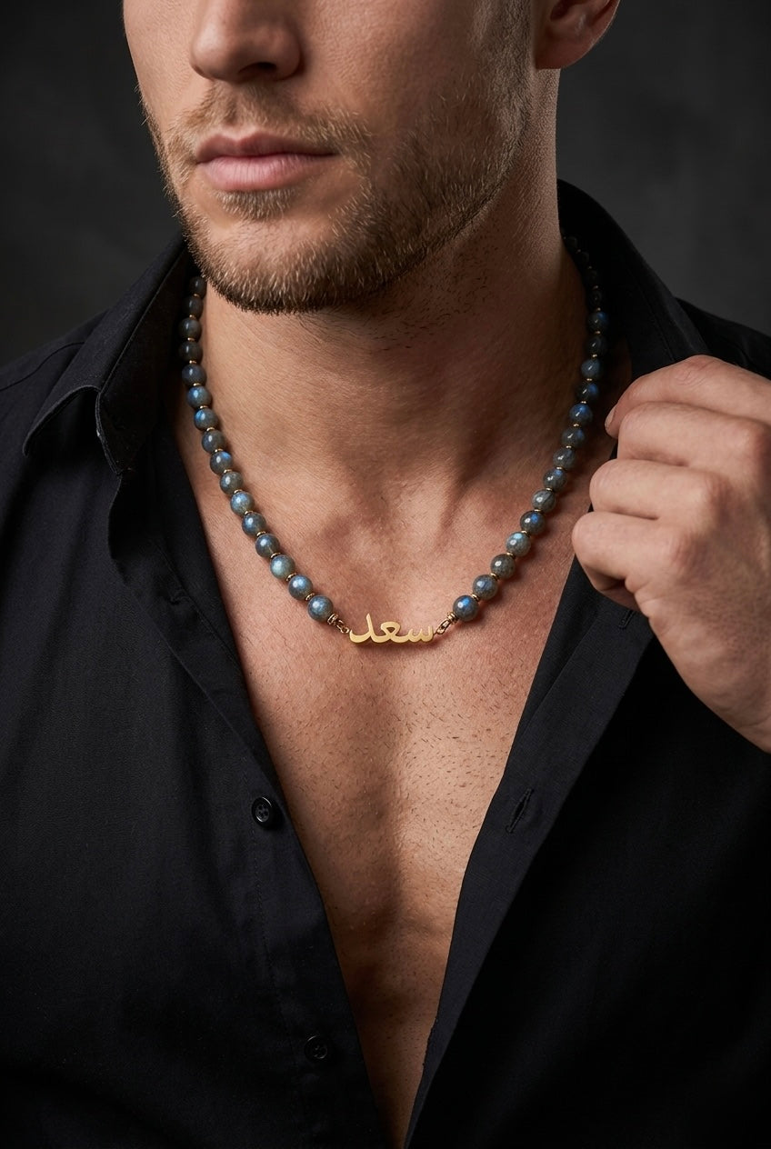 Man wearing a black shirt with a gold necklace featuring Arabic script against a dark background