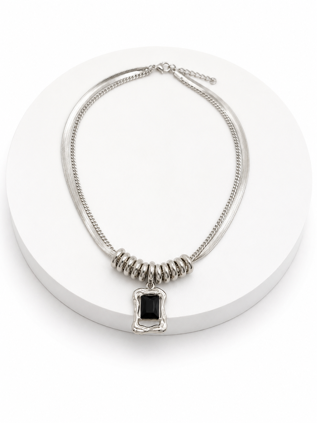 A silver-toned double-strand necklace with a textured central section and a rectangular black pendant, displayed on a white circular pedestal.