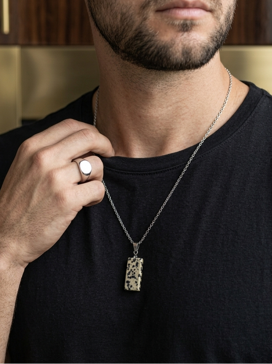 Man wearing a black shirt with a necklace and ring, against a blurred indoor background.